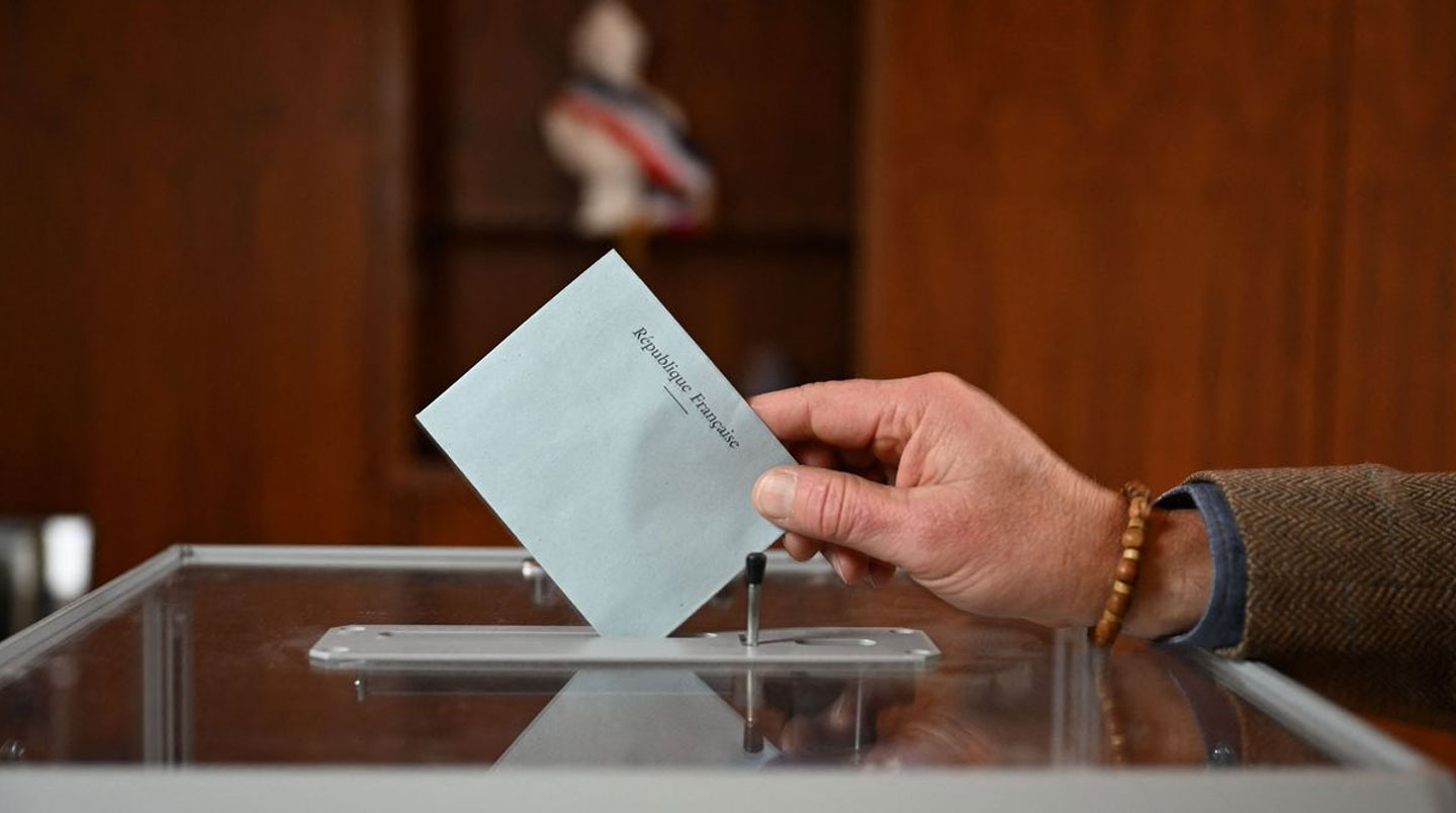 Bureau de vote CSE avec urne transparente en plexiglas, bulletins pliés à l'intérieur, pile de bulletins vierges, isoloirs en arrière-plan, affichette 'Bureau de vote - Élections CSE', salle vide
